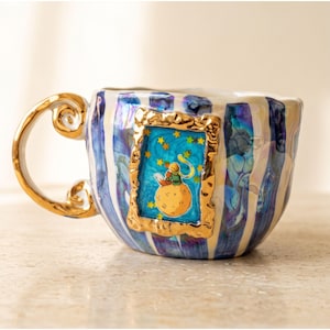 May include: A ceramic mug featuring vertical blue and white stripes with a gold-painted handle. A central rectangular gold frame displays an illustration of a small figure sitting on a planet with a fox, surrounded by yellow stars on a blue background.