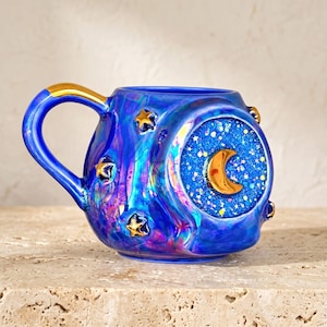 May include: A round, iridescent blue ceramic mug featuring a gold handle and gold star accents. The center of the mug displays a circular inset filled with blue glitter and a prominent, raised gold crescent moon shape in the middle of the glitter.