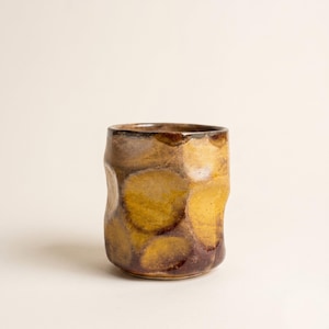 Puede incluir: Una taza de cerámica cilíndrica con una superficie irregular y abollada. El exterior presenta un esmalte moteado con parches superpuestos de color amarillo mostaza, marrón oscuro y tonos bronce apagados, creando un aspecto terroso y variado.