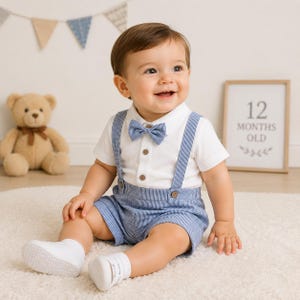 May include: A white short-sleeved collared shirt with brown buttons, paired with blue and white pinstriped shorts featuring matching suspenders and a small bow tie. The outfit is completed with white socks, creating a coordinated ensemble.