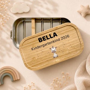 Peut inclure: Une bo&icirc;te &agrave; d&eacute;jeuner rectangulaire en m&eacute;tal avec un couvercle amovible en bambou. Le couvercle est grav&eacute; avec le nom BELLA, le texte Kindergarten 2026 et une petite illustration de z&egrave;bre. L'int&eacute;rieur du r&eacute;cipient en m&eacute;tal comporte un s&eacute;parateur.