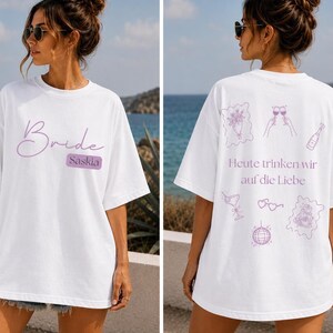 May include: A white t-shirt featuring the word Bride and the name Saskia in purple on the front. The back displays purple line art illustrations of a bouquet, wine glasses, a bottle, a cocktail, heart glasses, and a disco ball with the text Heute trinken wir auf die Liebe.