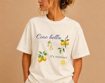 Puede incluir: Camiseta color crema con el texto "Ciao bella" y "it's summer". La camiseta presenta ilustraciones de acuarela de limones, una botella y un sol. La camiseta tiene mangas cortas y un corte holgado.