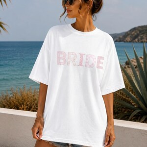 May include: A white oversized t-shirt featuring the word BRIDE printed across the chest in a light pink and white checkered pattern. The shirt has short sleeves, a crew neckline, and a loose fit, constructed from a solid white fabric.