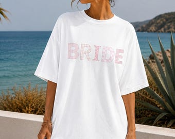 Puede incluir: Una camiseta blanca de corte oversize con la palabra BRIDE estampada en el pecho en un patrón de cuadros de color rosa claro y blanco. La prenda tiene mangas cortas, cuello redondo y un ajuste holgado, confeccionada en tela blanca.