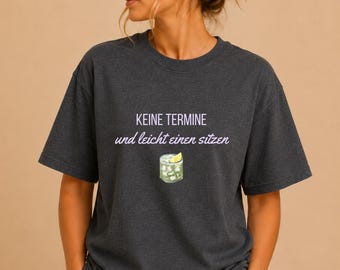 Puede incluir: Camiseta gris oscuro con el texto alemán "KEINE TERMINE und leicht einen sitzen" en morado, junto con un gráfico de un cóctel. La camiseta es de cuello redondo y tiene mangas cortas.