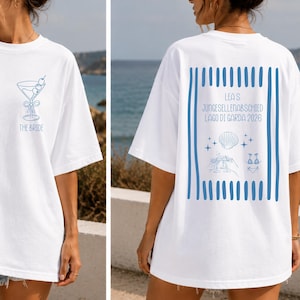 May include: A white short-sleeved t-shirt featuring a blue martini glass graphic and the text THE BRIDE on the front. The back displays blue vertical stripes, the text LEA'S JUNGESELLENABSCHIED LAGO DI GARDA 2026, and small shell and drink icons.