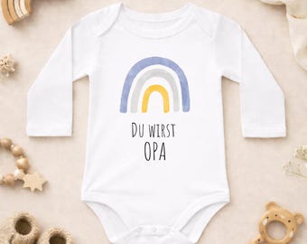 Puede incluir: Body de bebé blanco con mangas largas, con un arcoíris de acuarela en azul, gris y amarillo. El texto "DU WIRST OPA" está impreso debajo. La imagen incluye juguetes de madera y zapatos de bebé.