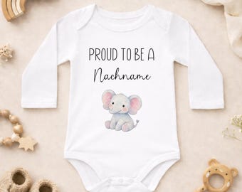 Puede incluir: Body de bebé blanco con mangas largas, con el texto "PROUD TO BE A Nachname" sobre una ilustración de acuarela de un elefante gris con orejas rosas. El body está rodeado de accesorios para bebés.