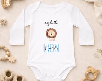 Puede incluir: Body de bebé blanco de manga larga con un dibujo de león y el nombre "Noah" en azul. El texto "my little" está encima de la ilustración del león. El body está rodeado de accesorios para bebés, incluyendo un arcoíris de madera y un mordedor.