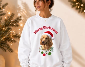 Puede incluir: Una sudadera blanca de manga larga con el gráfico de un perro de color dorado que lleva un gorro de Papá Noel rojo y blanco. El texto Merry Christmas está impreso en rojo sobre el perro, rodeado por una corona festiva con adornos rojos y verdes.