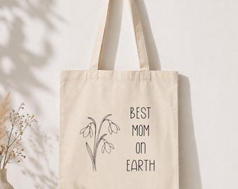 Puede incluir: Bolso tote de lona beige con el texto "BEST MOM ON EARTH" en negro. Un dibujo lineal negro de flores de campanillas está impreso debajo del texto. El bolso tote está colgado de una clavija de madera.