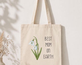 Puede incluir: Bolso tote de color crema con una ilustración floral y el texto "BEST MOM ON EARTH" en negro. La ilustración presenta flores de campanilla blancas con hojas verdes. El bolso tiene un diseño sencillo con dos asas.