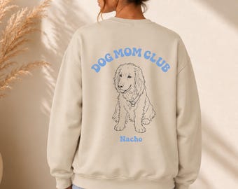Puede incluir: Una sudadera beige de cuello redondo con un gráfico azul claro en la espalda. El texto DOG MOM CLUB aparece en fuente arqueada sobre un dibujo lineal negro de un perro, con el nombre Nacho impreso en azul claro bajo la ilustración.