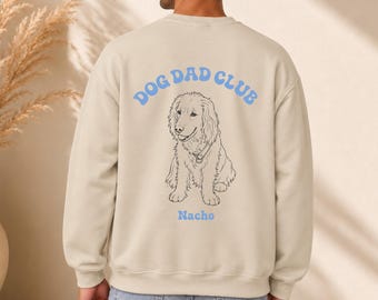 Puede incluir: Una sudadera beige de cuello redondo con el texto arqueado en azul claro que dice DOG DAD CLUB sobre una ilustración de línea negra de un perro de pelo largo. Debajo de la ilustración del perro, el nombre Nacho está impreso en azul claro.