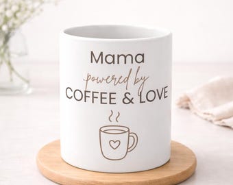 Puede incluir: Taza de cerámica blanca con el texto "Mama powered by COFFEE & LOVE" en escritura marrón. Un gráfico de taza de café con un corazón está debajo del texto. La taza está sobre un posavasos redondo de madera.