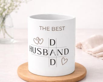 Puede incluir: Taza de cerámica blanca con el texto "THE BEST HUSBAND" en un diseño de cuadrícula, con símbolos de corazón y D. La taza está sobre un posavasos redondo de madera. Un jarrón con flores blancas está en el fondo.