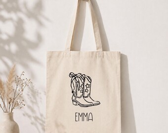 Puede incluir: Un bolso tote de color crema con un dibujo lineal negro de una bota de vaquero con detalles de estrellas y un lazo. El nombre "EMMA" está impreso en negro debajo de la ilustración de la bota. La bolsa cuelga de un gancho de madera.