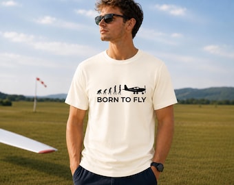 Puede incluir: Una camiseta de manga corta color crema con un estampado gráfico negro de la evolución humana progresando hacia un avión, acompañada del texto BORN TO FLY en una fuente sans-serif en negrita centrada en el pecho de la prenda.