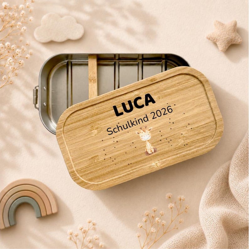 May include: An oval metal lunch box with a removable bamboo lid. The lid features the name LUCA, the text Schulkind 2026, and a small illustration of a giraffe. The interior of the lunch box is silver-coloured metal with a wooden divider.