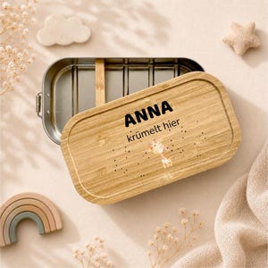 May include: A rectangular metal lunch box with a removable bamboo lid. The lid features the black text ANNA kr&uuml;melt hier and a small illustration of a giraffe. The interior of the metal container is divided by a single bamboo partition.