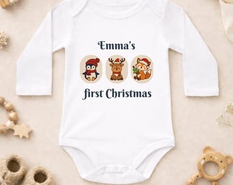 Puede incluir: Un body blanco de manga larga para bebé con el texto Emma's first Christmas en letras azul oscuro. El diseño incluye tres ilustraciones circulares de un pingüino, un reno y un zorro, cada uno con un gorro de Papá Noel y bufanda roja.