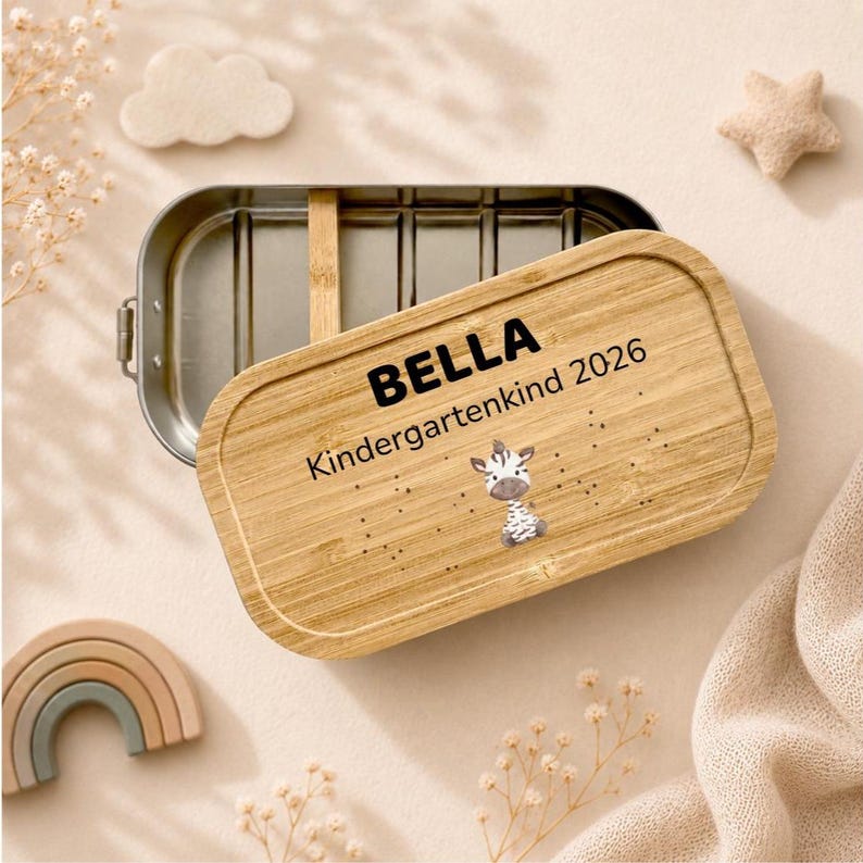 May include: A rectangular metal lunch box with a removable bamboo lid. The lid is engraved with the name BELLA, the text Kindergarten 2026, and a small illustration of a zebra. The interior of the metal container features a bamboo divider.
