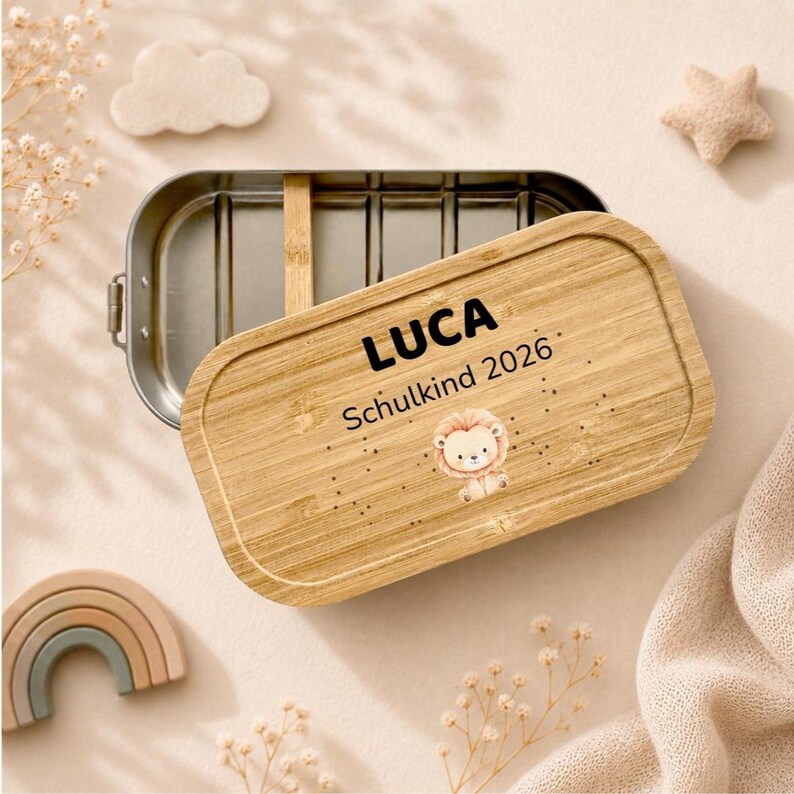May include: A rectangular stainless steel lunch box with a removable bamboo lid. The lid is engraved with the name LUCA, the text Schulkind 2026, and a small illustration of a lion cub. The container features a metal interior with a divider.