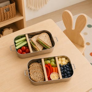 May include: Two rectangular stainless steel lunch boxes with metal side latches and removable wooden dividers. One container holds a sandwich, cucumber sticks, and cherry tomatoes. The other holds bread slices, grapes, bell peppers, carrots, dip, cheese, and blueberries.