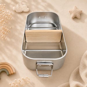 May include: A rectangular stainless steel lunch box with rounded corners, featuring a removable light-colored wooden divider in the center. The container has a metallic silver finish and a secure metal latch mechanism on the front exterior wall.