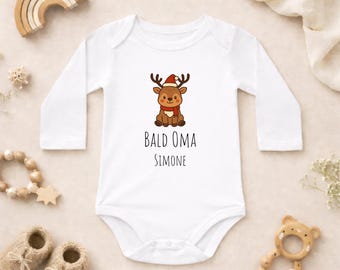 Puede incluir: Un body de bebé blanco de manga larga con un gráfico de un reno marrón que lleva un gorro de Papá Noel rojo y una bufanda roja. Debajo del reno, el texto dice "BALD OMA" seguido del nombre "SIMONE" en una fuente negra sencilla.