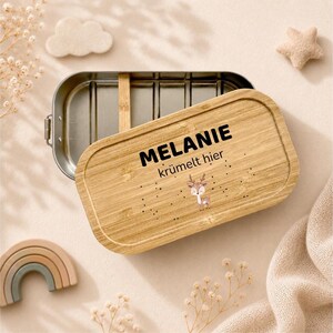 May include: A rectangular stainless steel lunch box with a removable bamboo lid. The lid features the printed text "MELANIE kr&uuml;melt hier" above a small illustration of a fawn, surrounded by scattered black dots on the light wood grain surface.