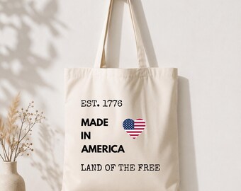 Puede incluir: Bolso tote de color crema con el texto "EST. 1776 MADE IN AMERICA LAND OF THE FREE". Un gráfico en forma de corazón de la bandera estadounidense está centrado en la bolsa. La bolsa está colgada de una clavija de madera contra una pared blanca.