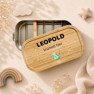 May include: A rectangular stainless steel lunch box with a removable bamboo lid. The lid features the black text LEOPOLD kr&uuml;melt hier, a small green dinosaur illustration, and scattered black dots. The interior has a single bamboo divider.