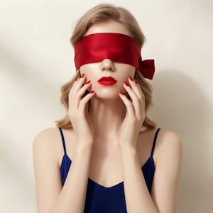 May include: A smooth red satin blindfold tied with a side bow, worn over the eyes. The fabric has a slight sheen and covers the upper face. The person wears a dark blue camisole with thin straps, and their fingernails are painted a matching red.