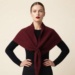 May include: A deep burgundy triangular shawl made of a soft, textured knit fabric. The shawl is draped over the shoulders and tied in a simple knot at the center front, resting over a plain black long-sleeved top and black trousers.