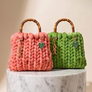 May include: Two chunky knit handbags, one coral and one green, with bamboo handles. Each bag features a charm with the letters "AKA" and a green leaf. The bags are displayed on a marble pedestal. The text "AmiraJoi legacy Collection" is at the bottom.