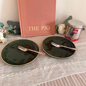 May include: Two oval, dark green plates with gold rims, each holding a silver fork. A cookbook titled "TALES and RECIPES from the KITCHEN GARDEN and beyond THE PIG" is in the background, along with a can of Illy coffee.