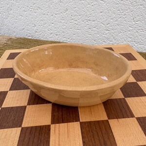 May include: A light brown, oval ceramic bowl with a slightly textured surface. The bowl has a simple, classic design, suitable for serving or display. The bowl is on a wooden cutting board.