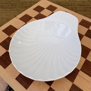 May include: A white, seashell-shaped dish with a textured, fan-like pattern. The dish is made of ceramic and has a smooth, glossy finish. The dish is placed on a wooden checkerboard.