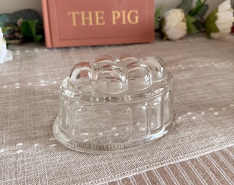 May include: A clear glass jelly mold with a rounded top and a decorative, ridged base. The mold sits on a striped, neutral-toned fabric. A book with the title "THE PIG" is in the background.