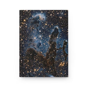 Op de afbeelding: Een hardcover notitieboek met een levendige afbeelding van de Adelaarsnevel, ook bekend als de Pilaren van de Schepping. De omslag toont een gedetailleerd beeld van kosmisch stof en gaswolken in tinten blauw, bruin en goud, met talloze sterren.