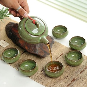 Puede incluir: Una tetera de cerámica verde vierte té en una taza a juego. La tetera y las tazas tienen un esmalte moteado. Cinco tazas adicionales están dispuestas alrededor de la tetera. El juego de té está sobre una estera tejida.