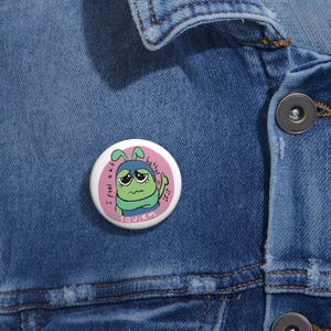 Pode incluir: Um pin de botão branco com uma ilustração de desenho animado de uma criatura verde triste com antenas azuis e o texto "I feel a bit better". O pin está sobre uma jaqueta jeans azul.