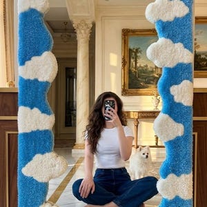May include: A large, rectangular mirror with a plush, cloud-shaped frame. The frame features a blue sky background with white cloud accents. The bottom edge has a green base with white and yellow flower accents.