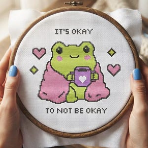May include: A circular wooden embroidery hoop containing white fabric with a cross-stitch design of a light green frog wrapped in a pink blanket, holding a purple mug with a white heart. The text reads IT'S OKAY TO NOT BE OKAY in black.