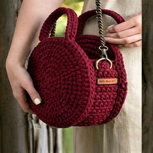 May include: A round, deep burgundy crochet handbag featuring a thick, textured knit pattern, two matching circular handles, and a metallic chain strap attached with gold-toned hardware. A small rectangular leather tag is attached to the side.
