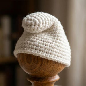 May include: A cream-colored, crocheted hat with a rounded top and a curled brim. The hat is displayed on a wooden stand, showcasing the texture of the yarn and the hat's shape. The hat appears to be handmade.