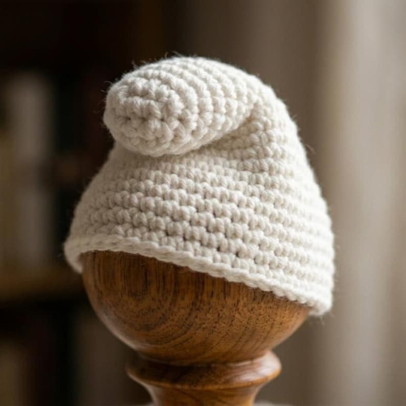 May include: A cream-colored, crocheted hat with a rounded top and a curled brim. The hat is displayed on a wooden stand, showcasing the texture of the yarn and the hat's shape. The hat appears to be handmade.