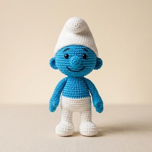 May include: A handmade crochet Smurf doll. The doll is blue with a white hat, white pants, and black eyes. The doll has a smiling expression and is standing upright. The crochet stitches are visible, giving the doll a textured appearance.